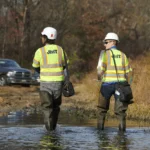 Two JMT employees wearing safety vests and hard hats wade through shallow water at a project site.